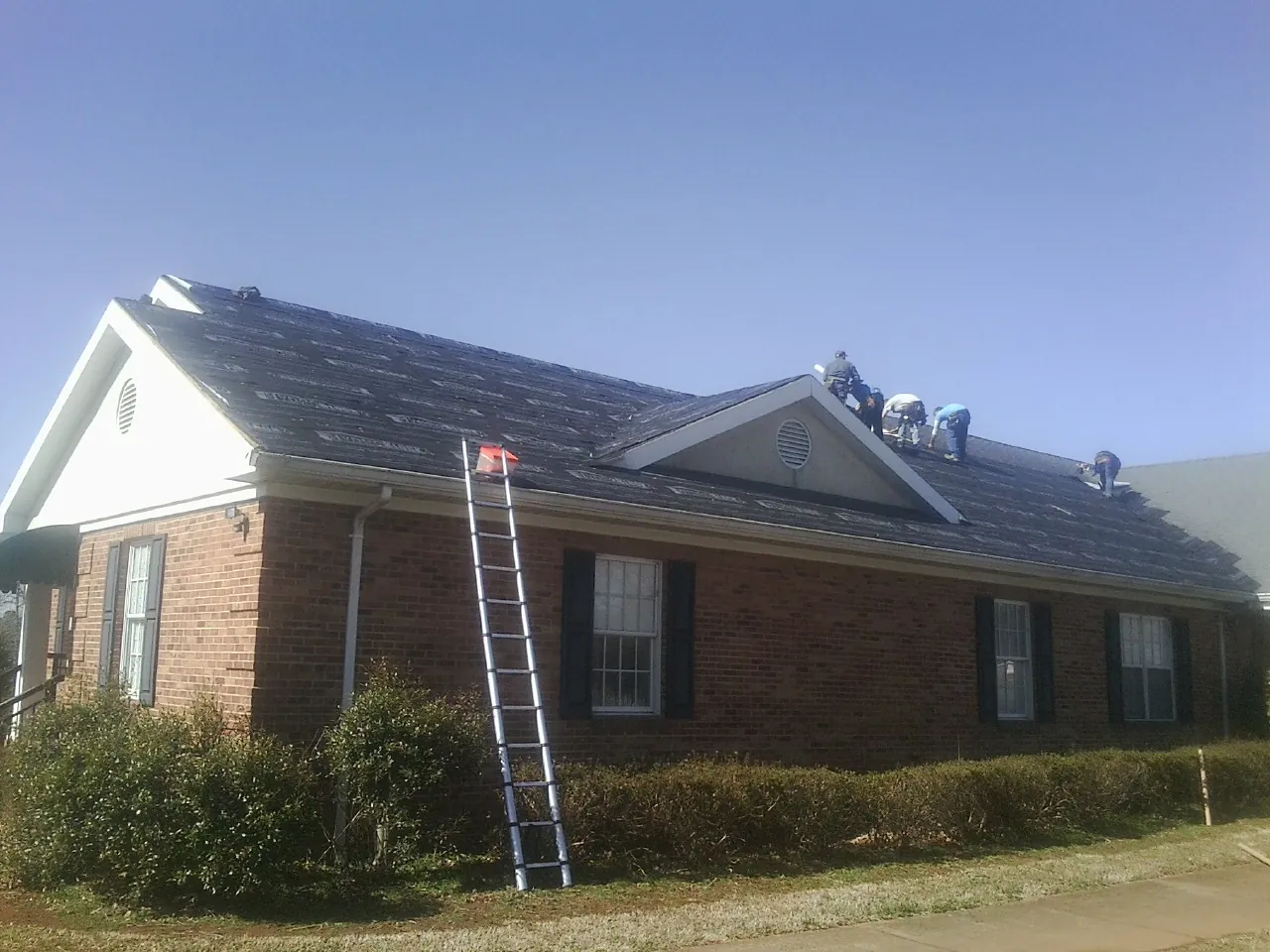 Crew tearing shingles off a brick ranch roof with a ladder set — Emergency Roof Repair work by our crew in Joanna
