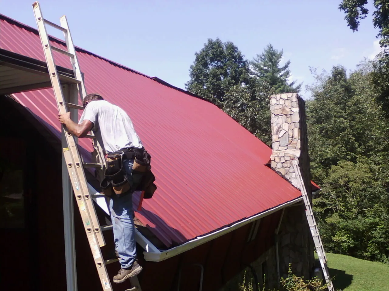 Roofer on a ladder beside a red metal roof and stone chimney — Soffit & Fascia Repair by Roofing Brothers in Joanna